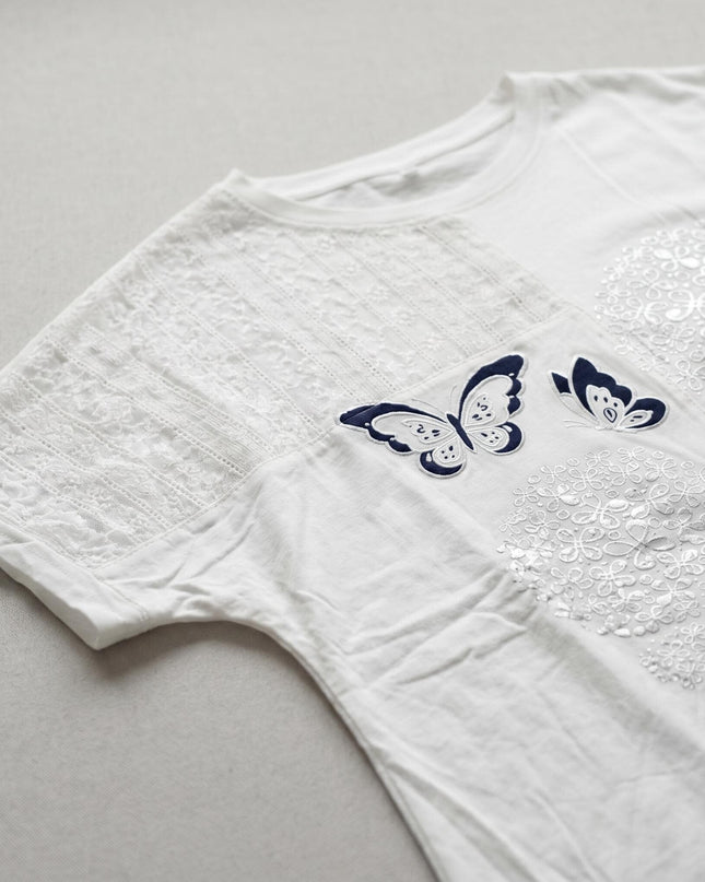 Детайл отблизо на бяла лятна памучна туника, показващ фин дантелен панел и изящна апликация с тъмносиня пеперуда и сребристи цветя.