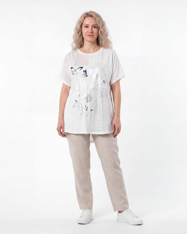 Бяла лятна памучна туника с флорална апликация и къс ръкав, представена от модел в цял ръст, съчетана със светъл панталон.