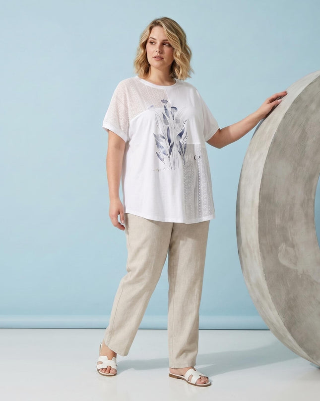 Бяла лятна туника големи размери с флорална апликация и къс ръкав, представена от модел. Кадър в цял ръст на светлосин фон с бежови панталони.