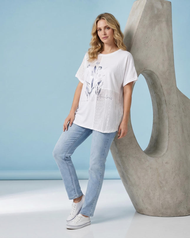 Бяла лятна туника големи размери с нежна флорална апликация. Кадър в цял ръст на модел със светли дънки, позиращ до скулптура на син фон.