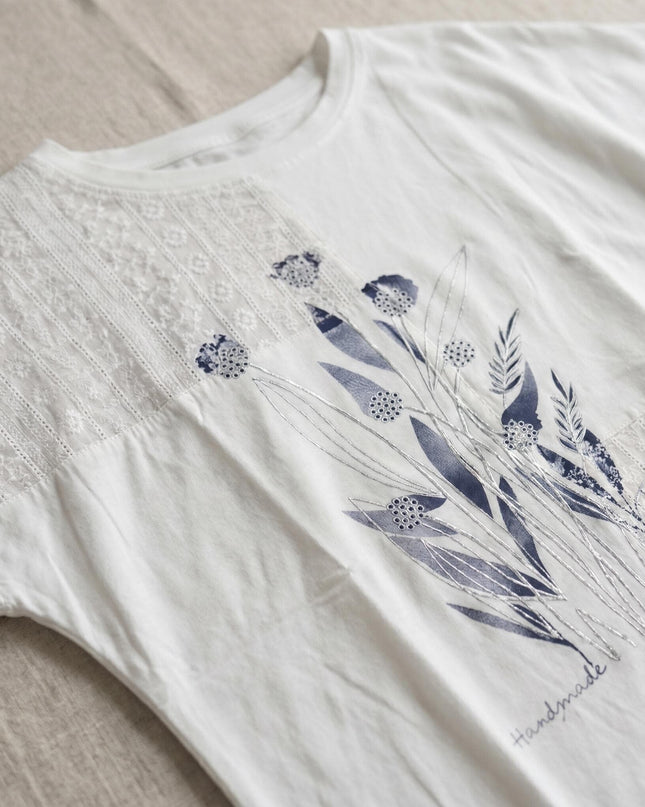 Детайл отблизо на бяла лятна туника големи размери, разкриващ красива флорална апликация и нежна дантела на рамото.