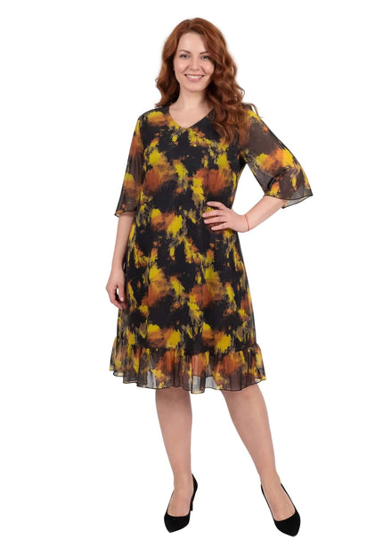 Черна официална шифонена рокля големи размери с жълт принт и волан на подгъва. Снимка в цял ръст, разкриваща елегантния силует и разкроените ефирни ръкави.