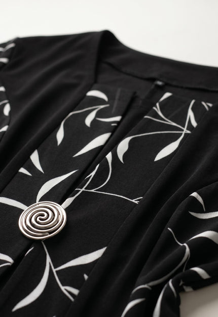 Детайл отблизо на черна официална рокля големи размери, показващ ефектен бял листен мотив и блестяща сребриста спираловидна брошка.