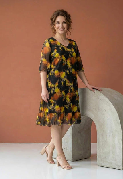 Черна официална шифонена рокля големи размери, заснета в цял ръст. Моделът се отличава с абстрактен жълто-оранжев принт и нежен волан на подгъва.