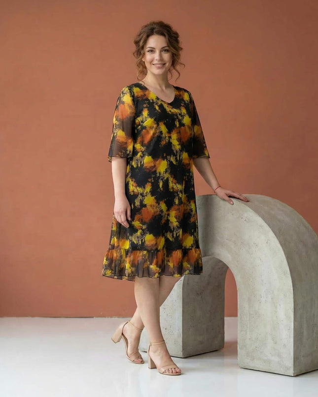 Черна официална шифонена рокля големи размери, заснета в цял ръст. Моделът се отличава с абстрактен жълто-оранжев принт и нежен волан на подгъва.