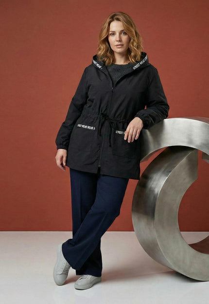 Черна разкроена макси ветровка с качулка и джобове, представена в цял ръст с акцент върху връзките на талията и декоративните надписи.