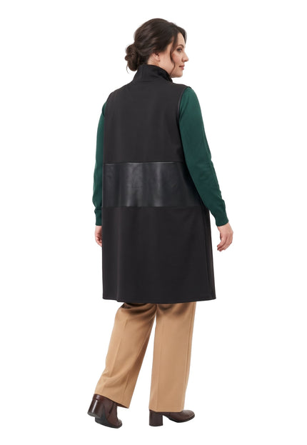 Жена, снимана отзад, е облечена с дълъг черен елек с широка текстурирана лента на гърба, зелен пуловер и бежов панталон на бял фон.