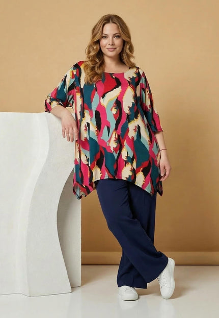 Дълга лятна макси туника в шарен абстрактен принт, представена в цял ръст върху модел, облегнат на бял декор.