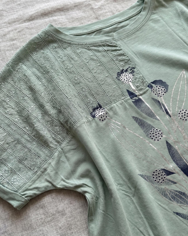 Детайл отблизо на ежедневна памучна туника големи размери в зелено, показващ текстурирани дантелени панели и ефектна апликация на цветя с блестящи акценти.
