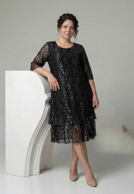 Официална черна макси рокля с ефектна дантела, разкроена кройка с етажиран подгъв и 3/4 дантелени ръкави. Моделът е в цял ръст, позиращ до бял пиедестал.