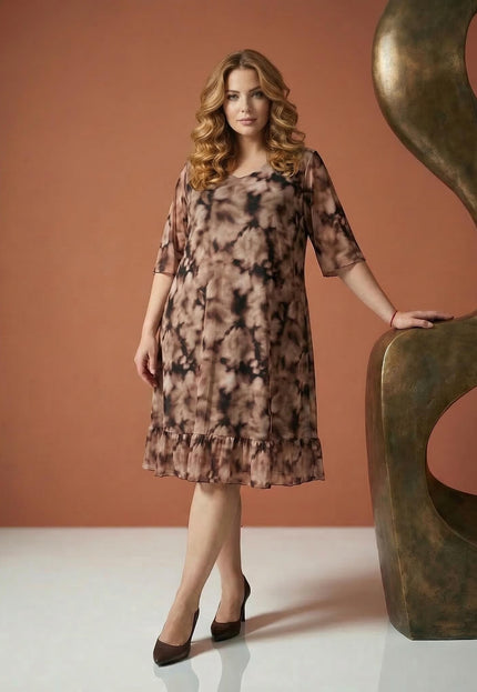 Официална макси рокля от ефирен шифон с преливащ десен в кафяво и подгъв с волан. Моделът позира в цял ръст до декоративна скулптура.