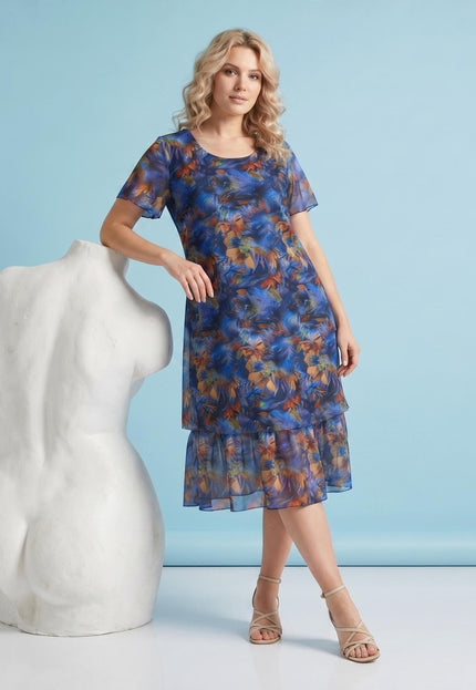 Цялостен изглед отпред на официална макси рокля с разкроен силует от пъстър шифон с цветен принт, къси ръкави и волани. Моделът е до скулптура на светлосин фон.