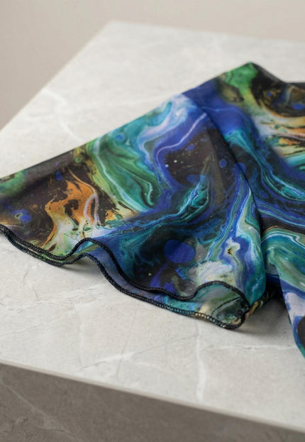 Детайл отблизо на официална шифонена рокля големи размери, показващ къс ръкав, преливащи шарени тонове и зашити ръбове, разположена върху светъл мраморен фон.