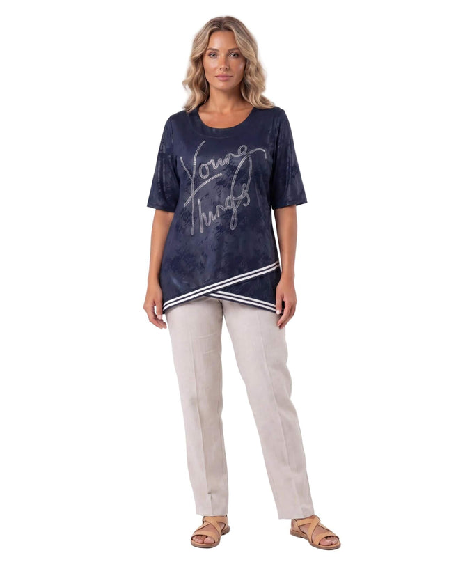 Тъмносиня туника големи размери с къс ръкав, блестящ надпис от камъчета и асиметричен раиран подгъв, представена в цял ръст.