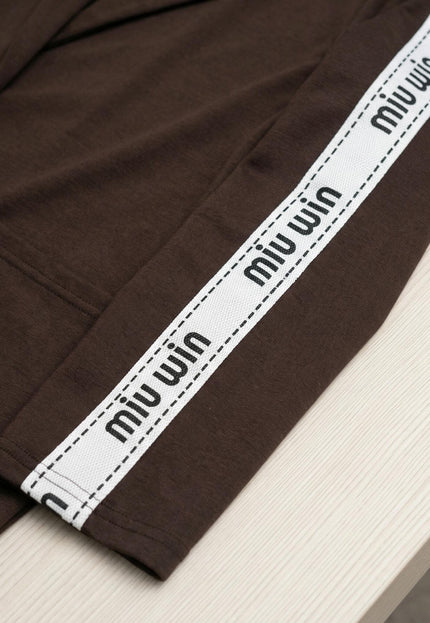 Детайлна снимка на ръкава на кафяв дамски комплект, акцентираща върху декоративната бяла лента с надпис miu win и текстурата на плата.