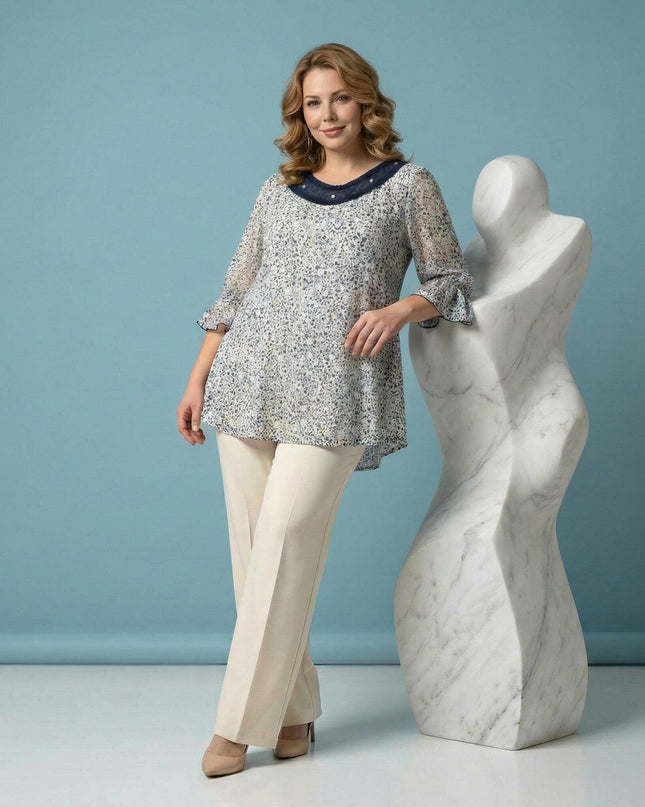 Стилна бяла макси туника с флорален принт и деколте с перли, представена от модел в цял ръст, опиращ се на бяла скулптура.