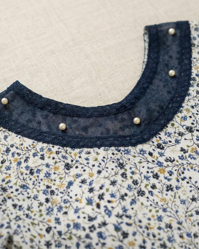 Детайл отблизо на деколтето на официална бяла макси туника на цветя, акцентирано с тъмносин тюл, дантелен кант и нежни перли.