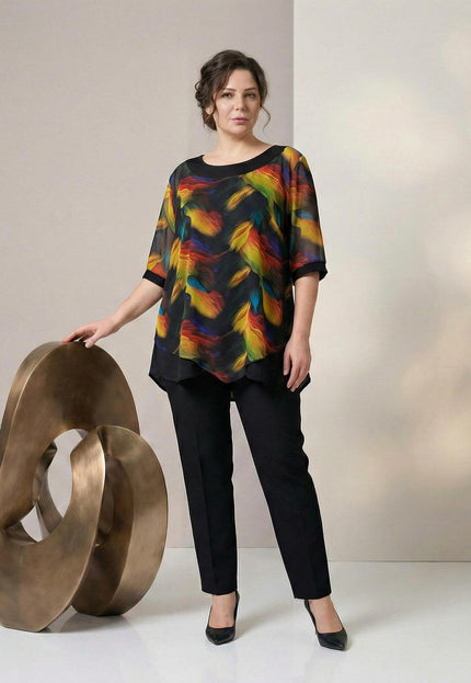 Елегантна макси туника с черна основа и ефирен пъстър шифон в асиметрична кройка, заснета в цял ръст върху модел до декоративна скулптура.