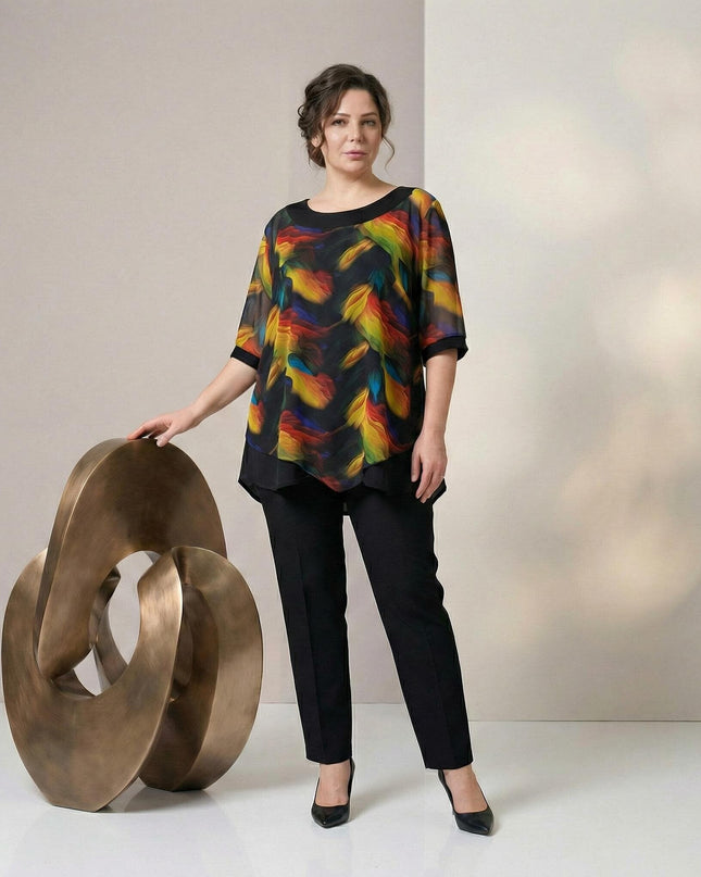 Елегантна макси туника с черна основа и ефирен пъстър шифон в асиметрична кройка, заснета в цял ръст върху модел до декоративна скулптура.