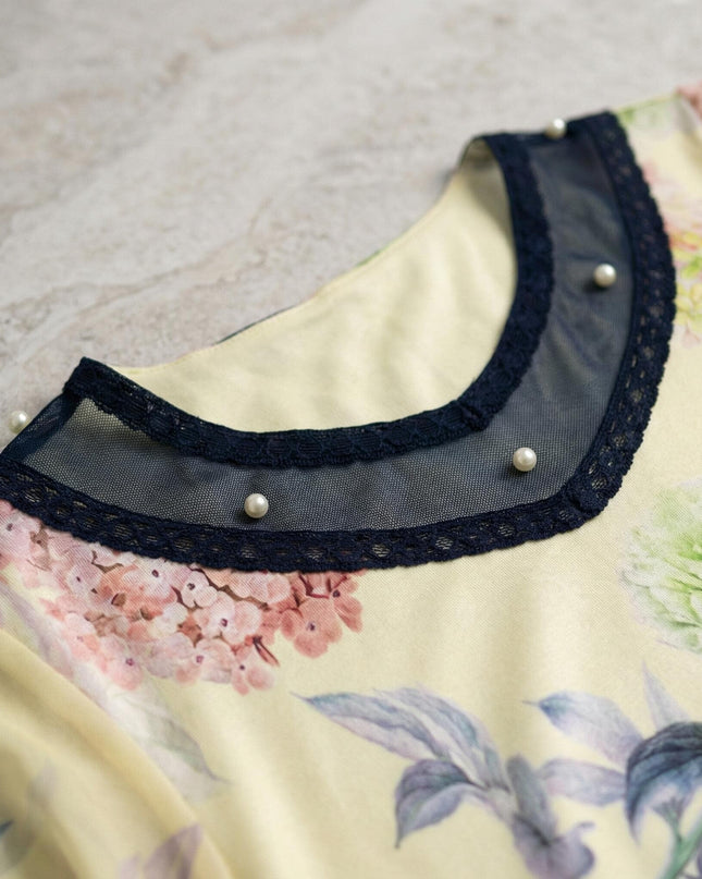 Детайл отблизо на деколтето на жълта макси туника на цветя. Акцентът е върху елегантната платка от тъмносин тюл с дантела и нежни бели перли.