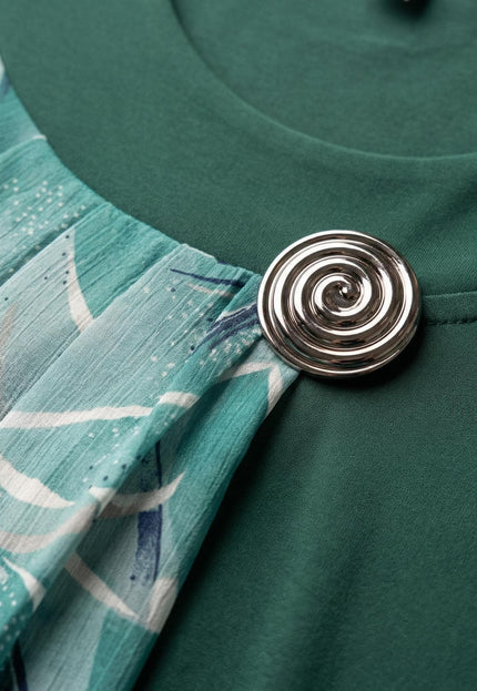 Детайлен поглед към сребристата брошка и набрания шифон на официална разкроена макси туника в масленозелено.