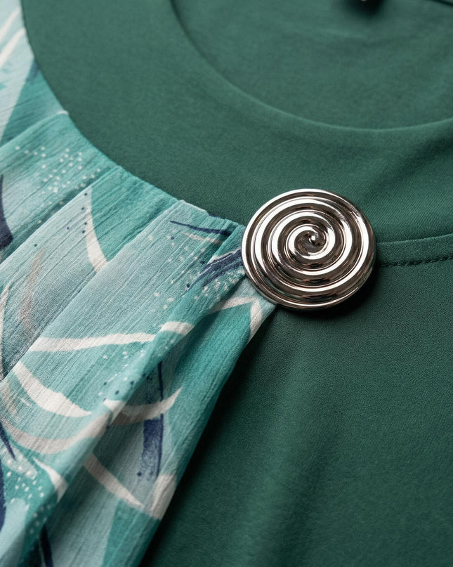 Детайлен поглед към сребристата брошка и набрания шифон на официална разкроена макси туника в масленозелено.