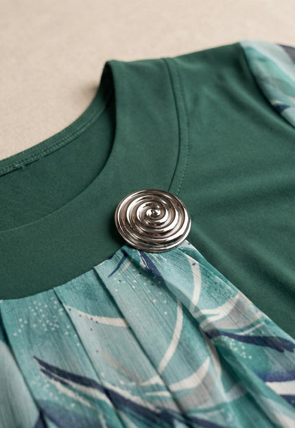 Детайлен кадър на деколтето на официална макси туника в масленозелено, акцентиращ върху елегантната сребриста брошка и набрания флорален шифон.