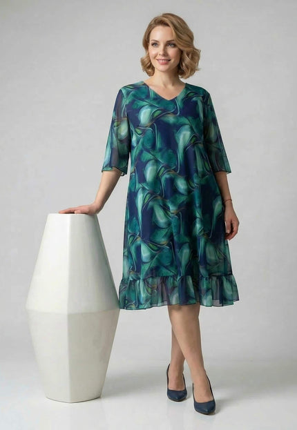 Официална шифонена рокля големи размери в преливащи сини и зелени тонове, цял ръст на бял фон, с V-образно деколте и волан.