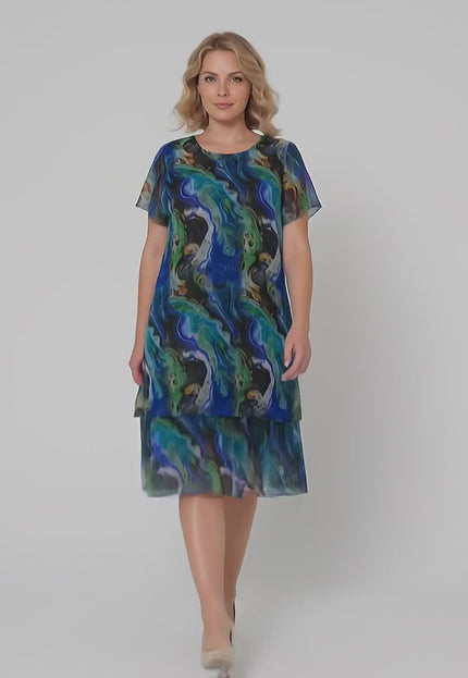 Плюс размер модел върви бавно към камерата в студийна среда на неутрален фон, носейки Официална шифонена рокля в преливащи тонове големи размери