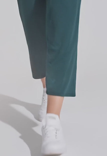 Плюс сайз модел, облечена с Еластичен тъмнозелен 7/8 панталон - Свободна кройка, върви бавно към камерата в студио с неутрален фон