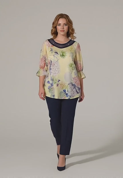 Макси модел върви бавно към камерата в студио на неутрален фон, облечена с Официална разкроена макси туника на цветя в жълто с перли и тюл