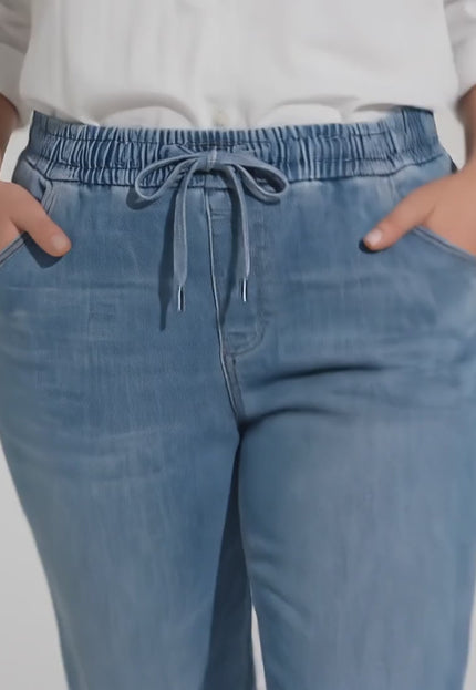 Студийно видео, в което плюс размер модел върви бавно към камерата на неутрален фон, облечена в продукта Сини макси дамски дънки с ластик и връзки от тенсел и памук