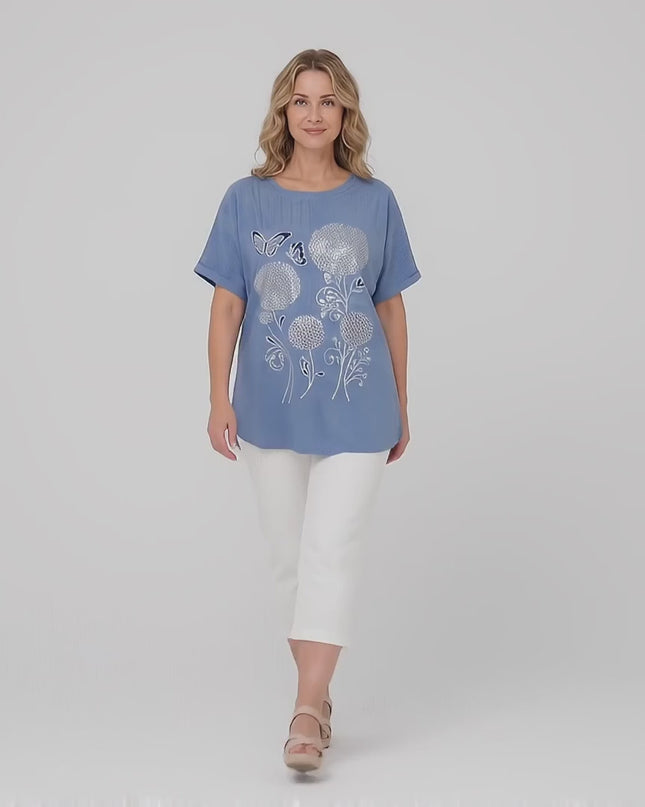 Студийно видео показващо как плюс сайз модел носи Дамска лятна синя туника големи размери с флорална апликация и върви бавно към камерата на неутрален фон