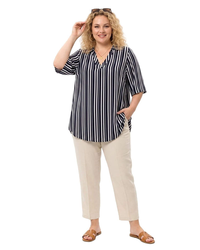 Разкроена дамска риза на тъмносиньо и бяло вертикално райе, съчетана с бежов панталон, представена в цял ръст на бял фон.