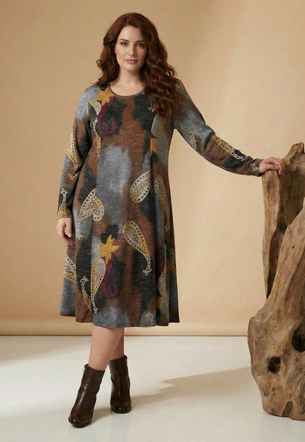 Дамска разкроена рокля в кафяво с едри пейсли и флорални мотиви, заснета в цял ръст. Елегантен модел с дълъг ръкав и А-силует, позиращ до дървена декорация.