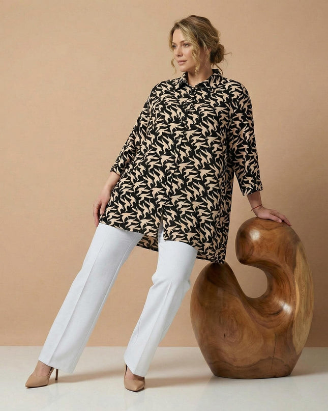 Цял ръст на жена, облечена в разкроена официална макси риза с десен в черно и бежово и бял панталон, подпряна на дървена скулптура.