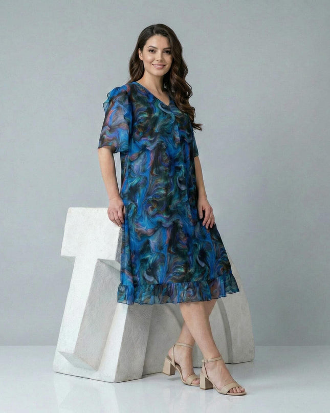 Шарена официална рокля големи размери с абстрактен принт и шифонени елементи, показана в цял ръст върху модел до бял декор.