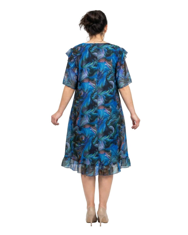 Шарена официална рокля големи размери в сини нюанси, заснета отзад. Вижда се свободният гръб на модела, ефирните шифонени ръкави и воланът в долната част.