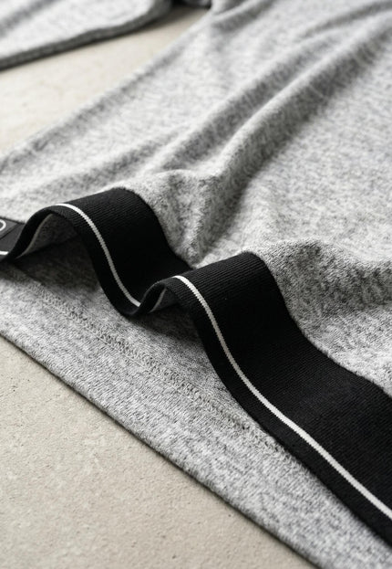 Детайл отблизо на подгъва на светлосива туника меланж с черна лента, бели кантове и лого нашивка, показващ структурата на меката вискозна материя.